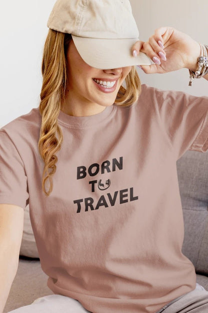 A woman wearing a Taupe t-shirt with the black printed text 'BORN TO TRAVEL' and a small smiley face graphic, paired with a beige baseball cap.