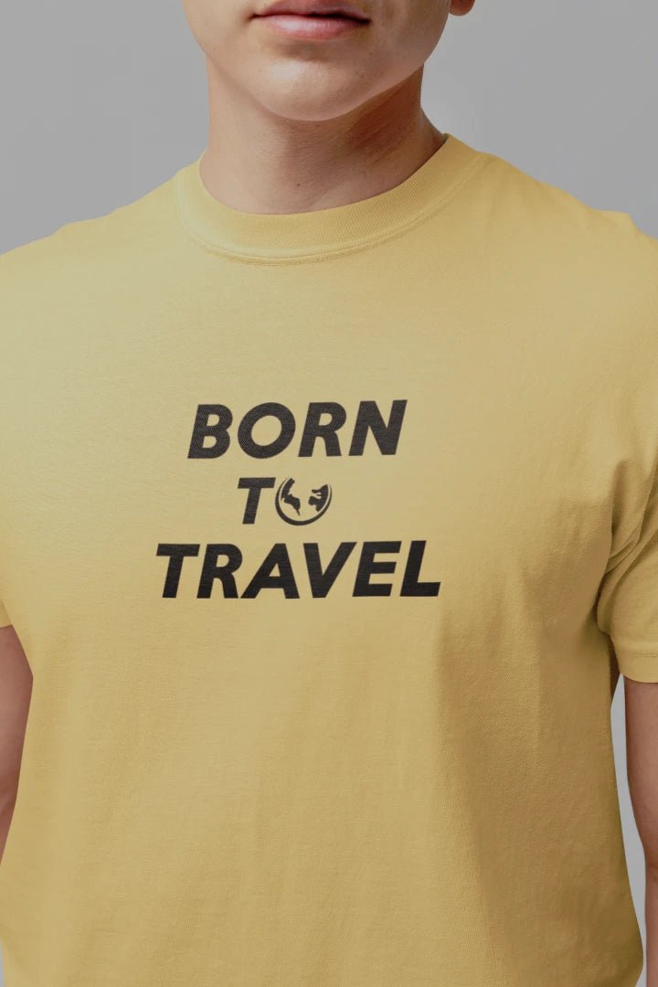 A close-up of a person wearing a Warm Sand crewneck t-shirt with the black printed text 'BORN TO TRAVEL' on the chest, set against a solid gray background.