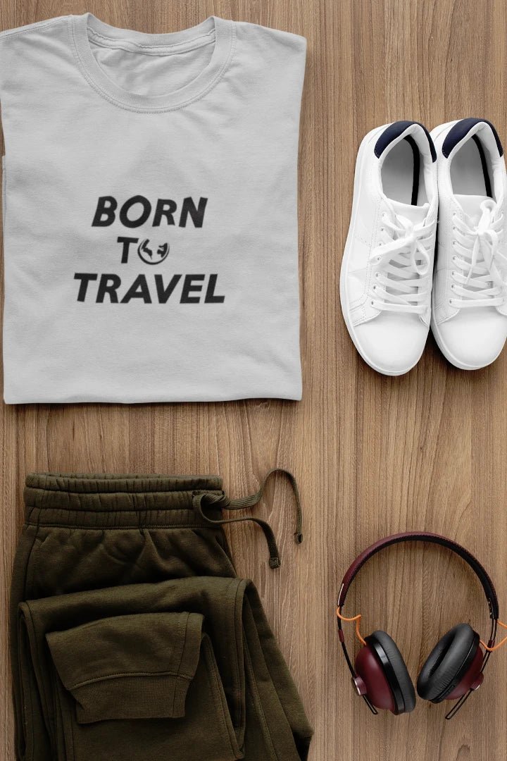 A styled flat-lay outfit on a wood background featuring a Heather Grey t-shirt with the black text 'BORN TO TRAVEL', olive green sweatpants, white sneakers, and black-and-burgundy over-ear headphones.