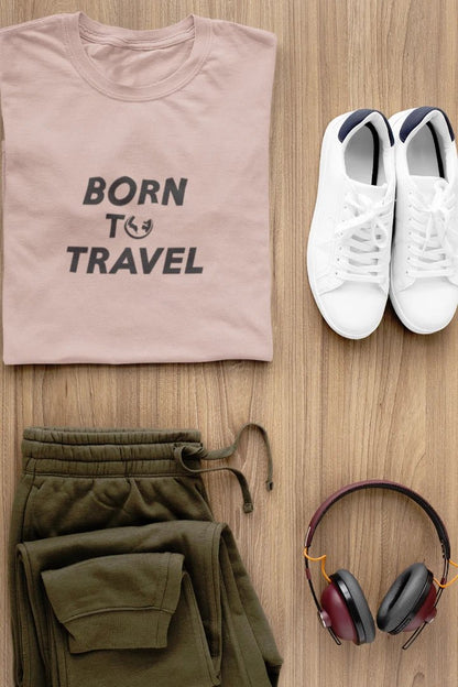 A styled flat-lay of a Taupe t-shirt with the text 'BORN TO TRAVEL' in dark gray, paired with olive green sweatpants, white sneakers, and over-ear headphones on a light wood background.