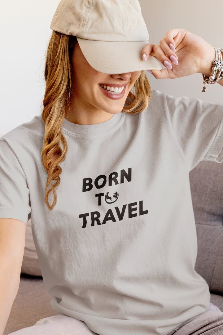 A woman wearing a Heather Grey t-shirt with the black printed text 'BORN TO TRAVEL' and a small smiley face graphic, paired with a beige baseball cap.