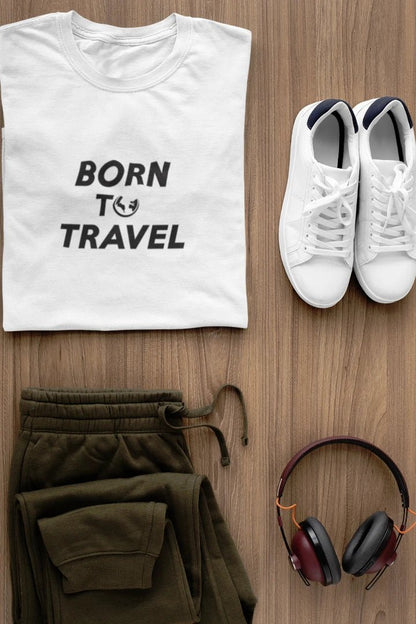 A styled flat-lay featuring a Pure White t-shirt with the black text 'BORN TO TRAVEL' and a small globe graphic, paired with olive green sweatpants, white sneakers, and black-and-burgundy headphones on a light brown wood background.