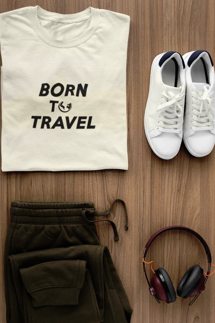 A styled flat-lay featuring a Off White t-shirt with the black text 'BORN TO TRAVEL' and a small globe graphic, paired with olive green sweatpants, white sneakers, and black-and-burgundy headphones on a light brown wood background.