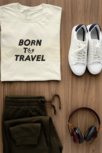 A styled flat-lay featuring a Off White t-shirt with the black text 'BORN TO TRAVEL' and a small globe graphic, paired with olive green sweatpants, white sneakers, and black-and-burgundy headphones on a light brown wood background.