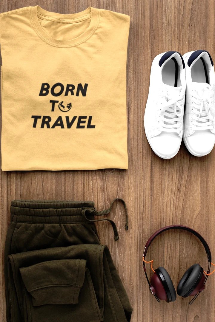 A styled flat-lay outfit on a wood background featuring a Warm Sand t-shirt with the black text 'BORN TO TRAVEL', olive green sweatpants, white sneakers, and black and burgundy headphones.