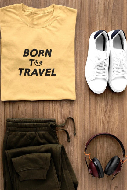 A styled flat-lay outfit on a wood background featuring a Warm Sand t-shirt with the black text 'BORN TO TRAVEL', olive green sweatpants, white sneakers, and black and burgundy headphones.