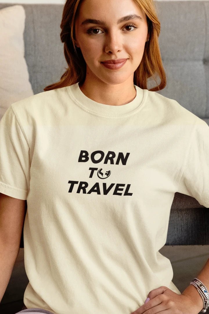 A woman wearing a Off White t-shirt with the black printed text 'BORN TO TRAVEL' and a small globe icon, posed in front of a gray couch.