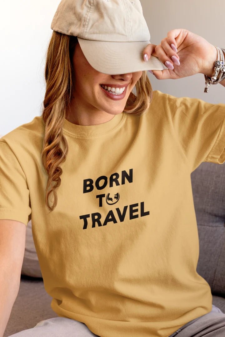 A woman wearing a Warm Sand t-shirt with the black printed text 'BORN TO TRAVEL' and a small globe graphic, paired with a beige baseball cap.