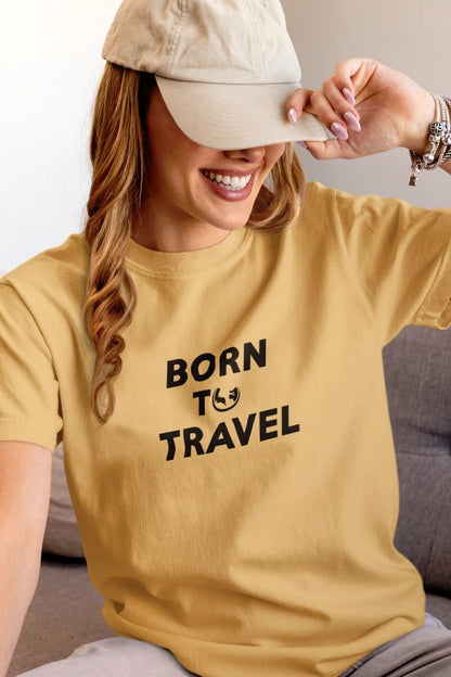 A woman wearing a Warm Sand t-shirt with the black printed text 'BORN TO TRAVEL' and a small globe graphic, paired with a beige baseball cap.