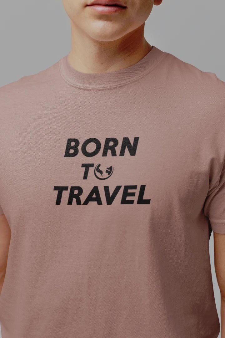A close-up of a person wearing a Taupe t-shirt with the black printed text 'BORN TO TRAVEL' on the chest, set against a solid gray background.