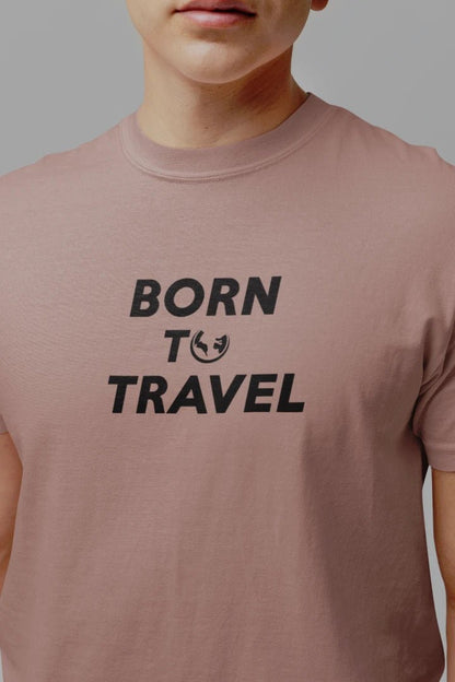 A close-up of a person wearing a Taupe t-shirt with the black printed text 'BORN TO TRAVEL' on the chest, set against a solid gray background.