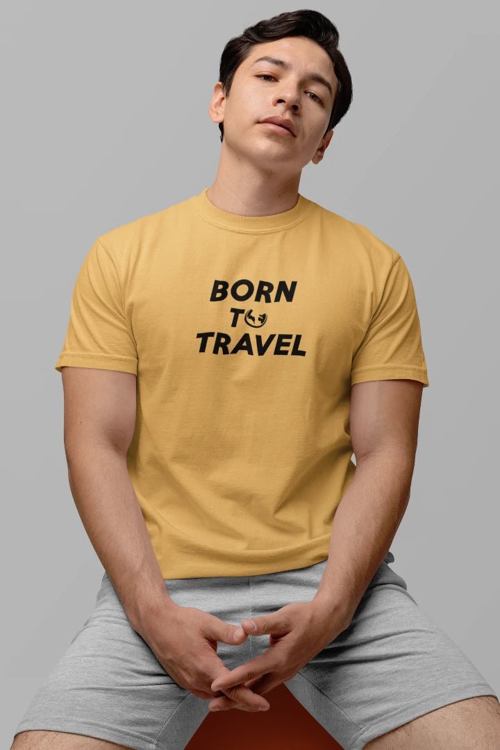 A man wearing a Warm Sand t-shirt with the black text 'BORN TO TRAVEL' printed on the chest, paired with light gray shorts, seated against a solid gray background.
