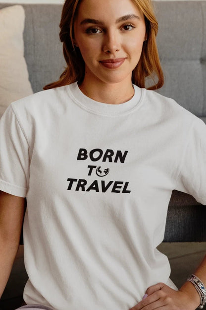 A woman wearing a Pure White crewneck t-shirt with the black printed text 'BORN TO TRAVEL' and a small globe icon, sitting in front of a gray tufted sofa.