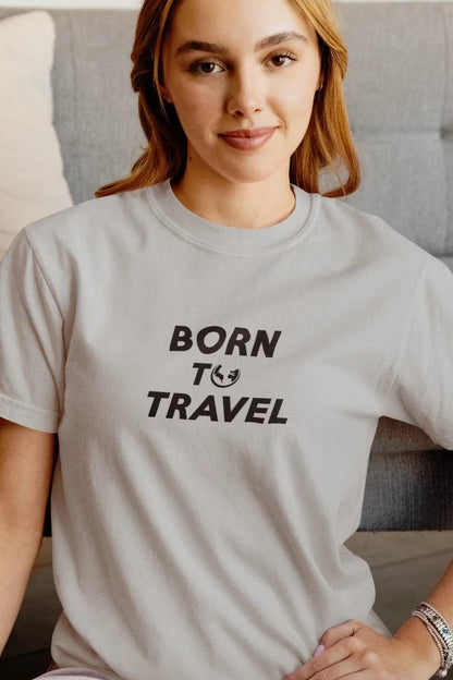 A woman wearing a Heather Grey crewneck t-shirt with the black printed text 'BORN TO TRAVEL' and a small globe icon, sitting in front of a gray tufted sofa.