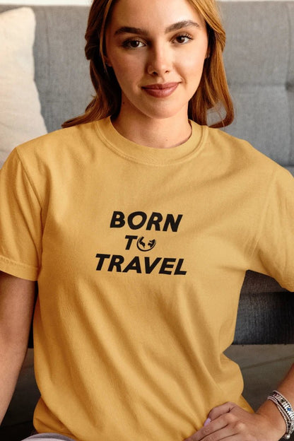 A woman wearing a Warm Sand t-shirt with the text 'BORN TO TRAVEL' printed in black on the chest, sitting in front of a gray couch.
