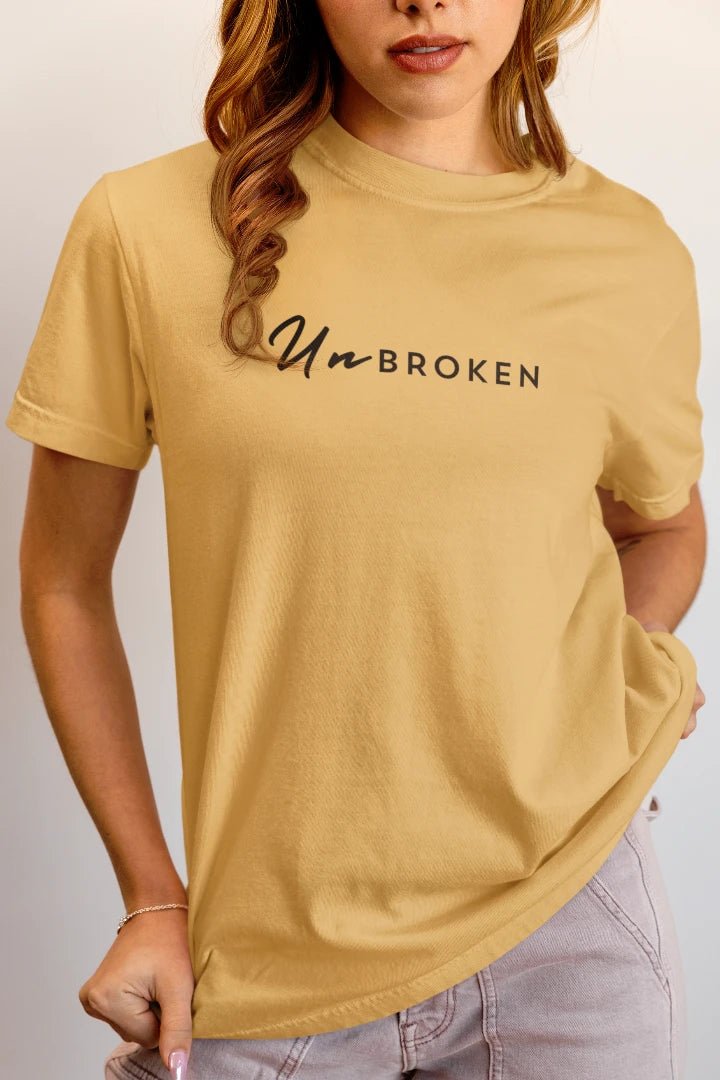 A woman wearing a beige t-shirt with the text 'Un-BROKEN' printed across the chest in black script and sans-serif fonts, paired with light gray jeans.