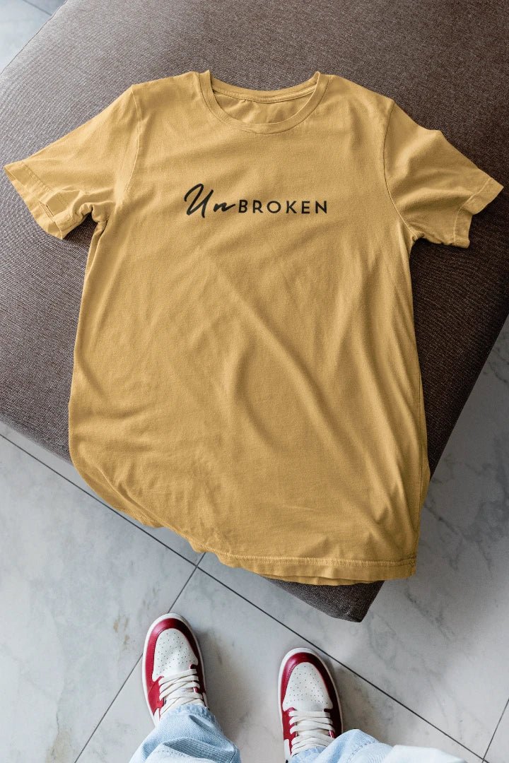 A beige t-shirt with the black printed text 'UnBROKEN' across the chest, laid flat on a brown upholstered ottoman with a person's feet in red and white sneakers visible at the bottom.