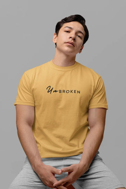 A man wearing a beige t-shirt with the text 'UnBROKEN' printed in black across the chest, paired with light gray sweatpants, set against a solid gray background.