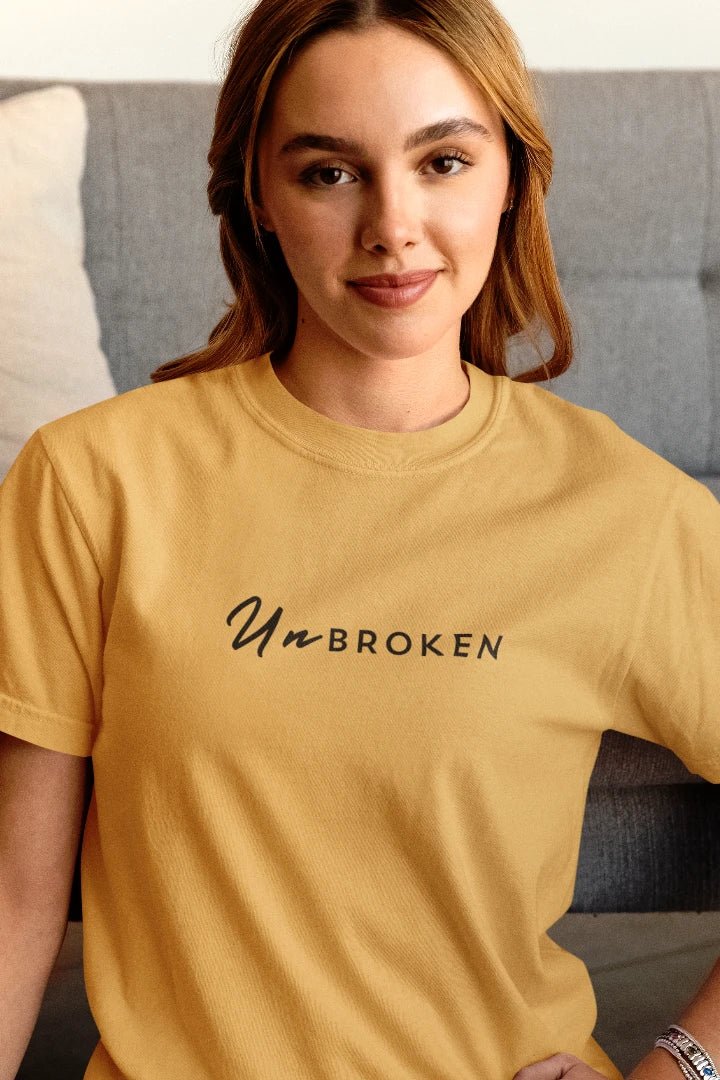 A woman wearing a beige t-shirt with the black printed text 'UnBROKEN' across the chest, posed in front of a gray couch.