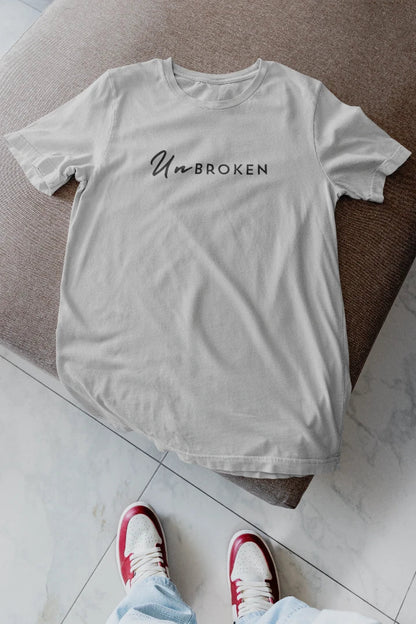 A grey  t-shirt with the text 'UnBROKEN' printed in black across the chest, laid flat on a brown upholstered ottoman with a person's feet in red and white sneakers visible at the bottom.