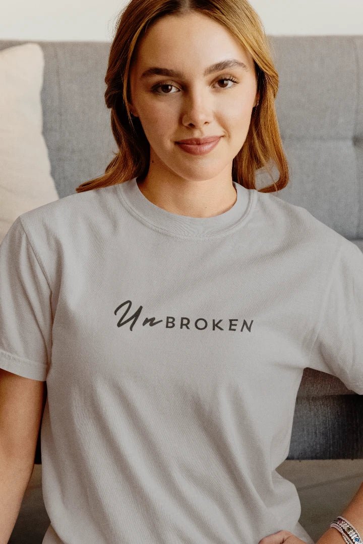 A woman wearing a grey crewneck t-shirt with the text 'Un BROKEN' printed in black across the chest, posed in front of a gray couch.