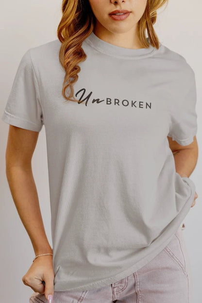 A woman wearing a grey crewneck t-shirt with the text 'Un-BROKEN' printed across the chest in black script and block lettering, paired with light pink jeans.
