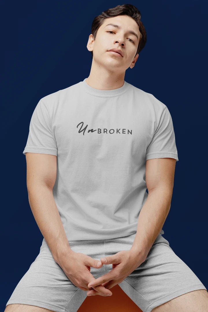 A man wearing a light grey crewneck t-shirt with the text 'UnBROKEN' printed in black across the chest, paired with matching light gray shorts, posed against a dark navy blue background.