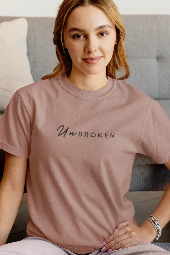 A woman wearing a mushroom t-shirt with the text 'Un BROKEN' printed in black across the chest, sitting in front of a gray tufted sofa.
