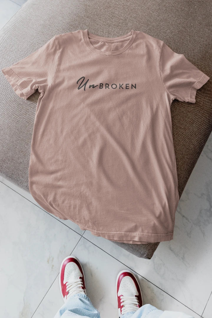A mushroom t-shirt with 'UnbROKEN' printed in black across the chest, laid flat on a gray upholstered bench with a person's feet in red and white sneakers visible at the bottom.