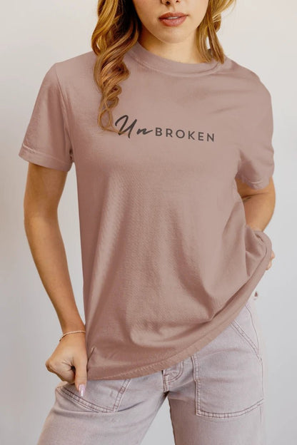 A woman wearing a mushroom t-shirt with the text 'Un-BROKEN' printed across the chest, paired with light-wash jeans against a plain light gray background.