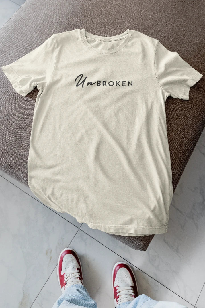 A off White t-shirt with the text 'UnBROKEN' printed across the chest, laid flat on a brown upholstered ottoman with a person's feet in red and white sneakers visible at the bottom.
