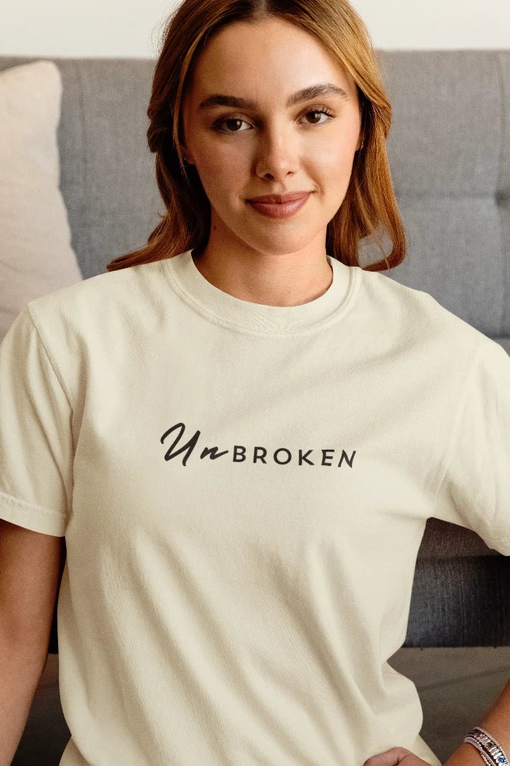 A woman wearing a off White t-shirt with the text 'Un BROKEN' printed in black across the chest, posed in front of a gray couch.