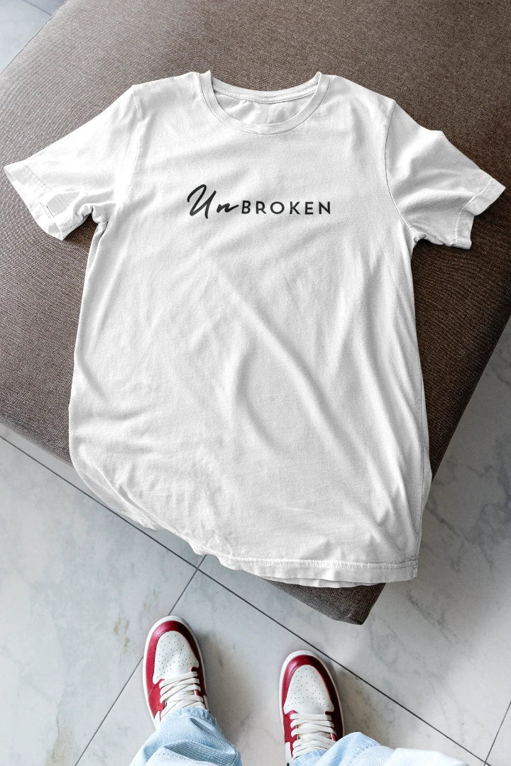 A white short-sleeve T-shirt with the text 'UnBROKEN' printed across the chest in black, laid flat on a brown upholstered bench. The bottom of the frame shows a person's feet wearing red and white sneakers and light blue jeans on a light gray marble floor.