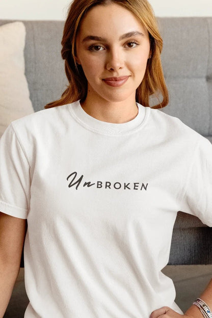 A woman wearing a white crewneck t-shirt with the black printed text 'Un BROKEN' across the chest, posed in front of a gray sofa.