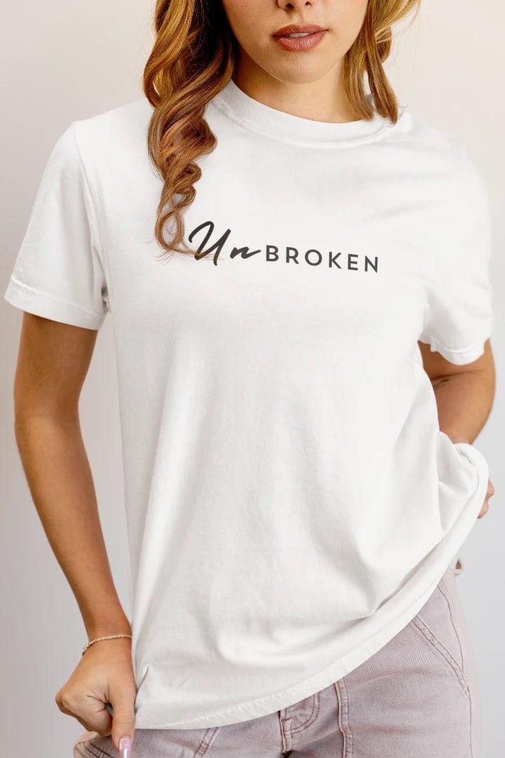 A woman wearing a white short-sleeve crewneck t-shirt with the text 'Un-BROKEN' printed across the chest in black script and block lettering, paired with light gray jeans against a plain light background.