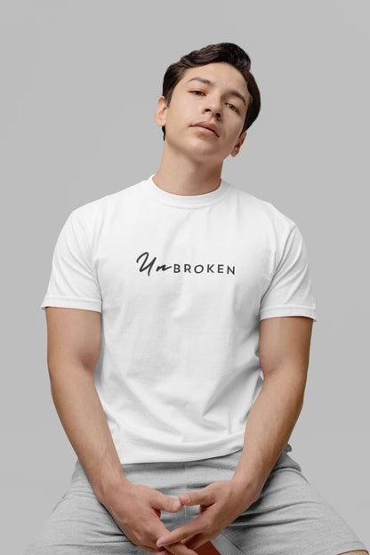 A man wearing a white crewneck t-shirt with the black printed text 'UnbROKEN' on the chest, paired with light gray sweatpants, set against a solid gray background.