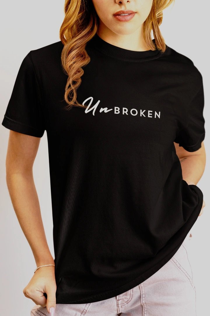 A woman wearing a black short-sleeve t-shirt with the word 'UnbROKEN' printed in white across the chest, paired with light pink jeans against a plain light gray background.