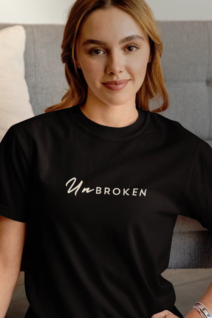 A woman wearing a black crewneck t-shirt with the words 'UnBROKEN' printed in white across the chest, posed in front of a gray couch.