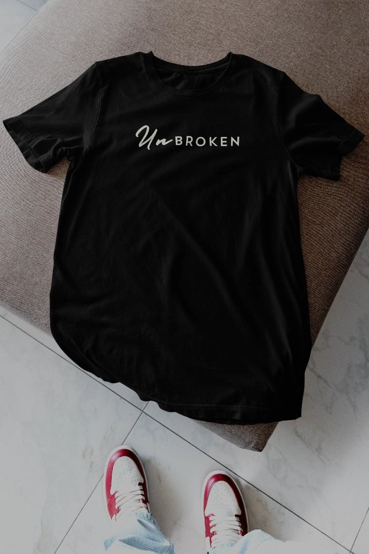 A black short-sleeve t-shirt with the text 'UnBROKEN' printed in white across the chest, laid flat on a brown upholstered bench with a person's feet in red and white sneakers visible at the bottom.