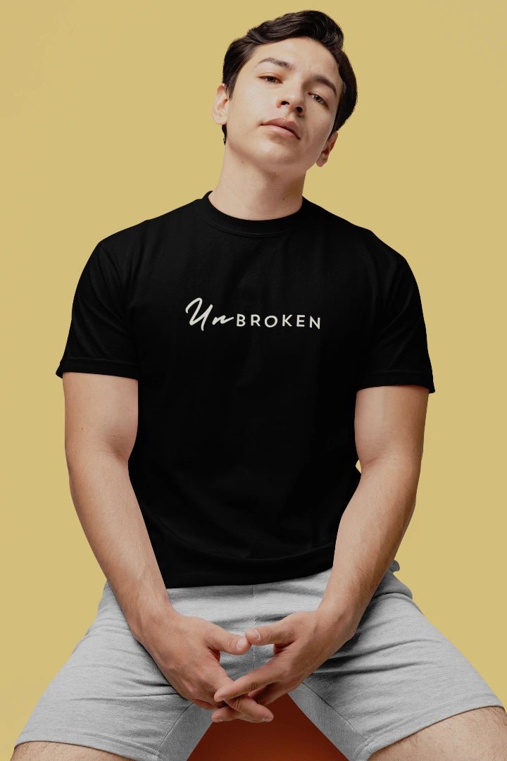 A man wearing a black crewneck t-shirt with the white text 'UnbROKEN' printed across the chest, paired with light gray shorts, seated against a solid mustard yellow background.
