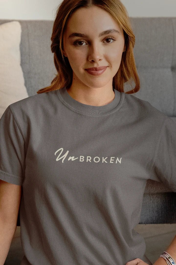 A woman wearing a Charcoal crewneck t-shirt with the words 'Un BROKEN' printed in white across the chest, posed in front of a gray couch.