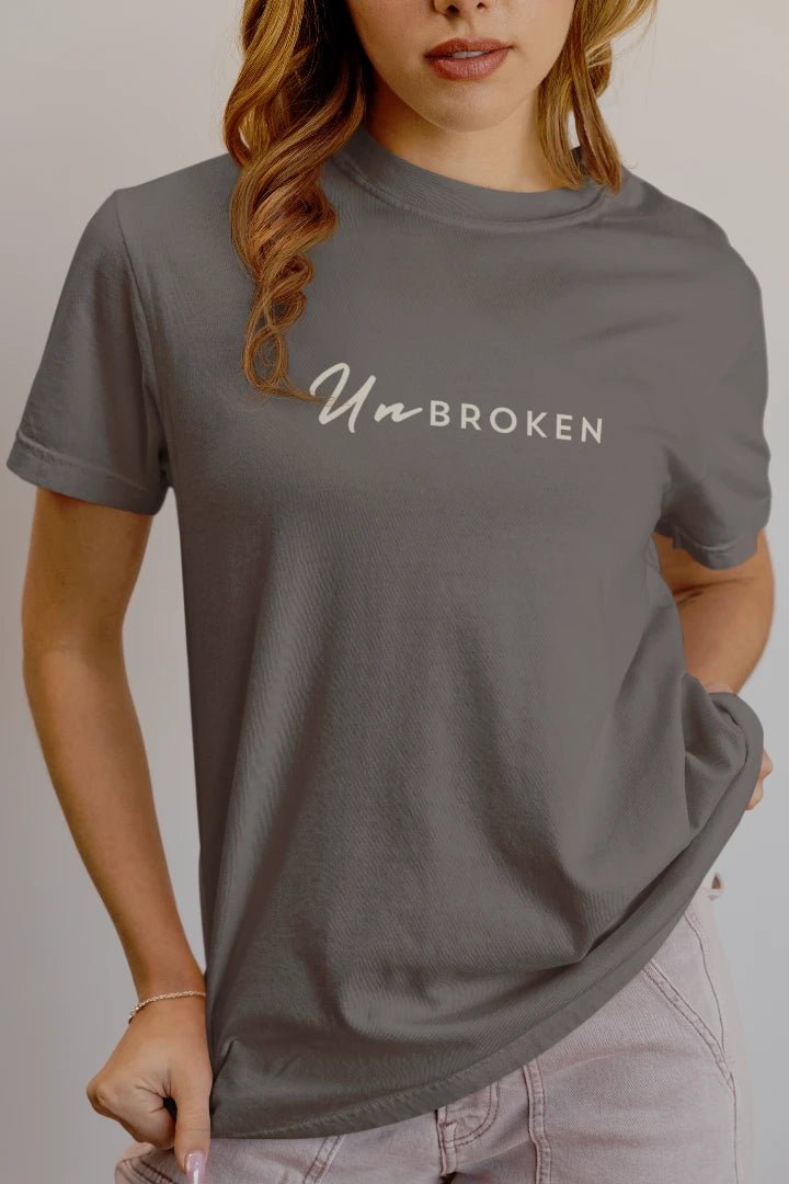 A woman wearing a Charcoal  crewneck t-shirt with the text 'UnbROKEN' printed in white across the chest, paired with light purple jeans against a plain light gray background.