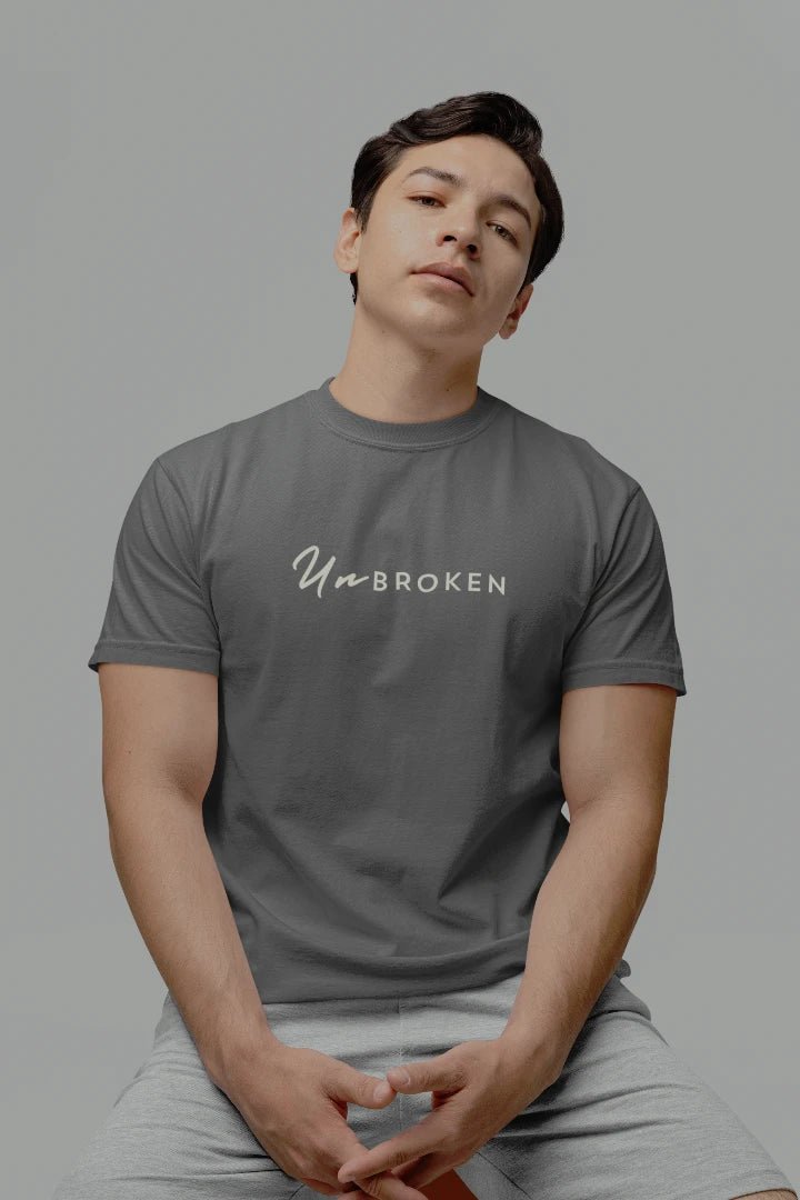 A man wearing a Charcoal  crewneck t-shirt with the words 'UnbROKEN' printed in white script across the chest, paired with light gray sweatpants, set against a solid gray studio background.