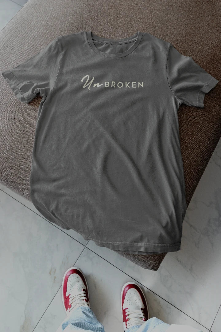 A Charcoal crewneck t-shirt with 'UnBROKEN' printed in white across the chest, laid flat on a brown upholstered ottoman with a person's feet in red and white sneakers visible at the bottom.