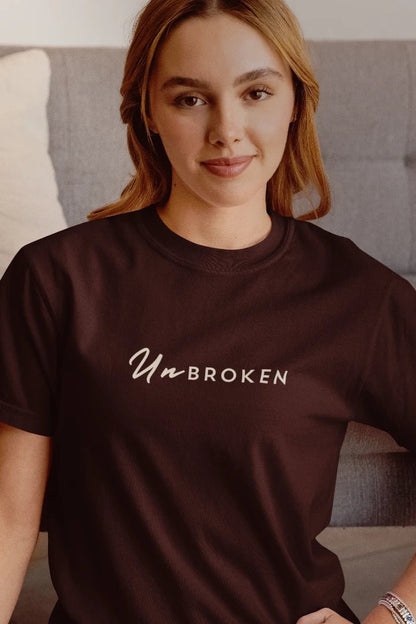 A woman wearing a coffee brown crewneck t-shirt with the words 'UnBROKEN' printed in white across the chest, posed in front of a gray sofa.