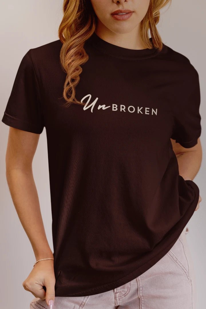 A woman wearing a coffee brown short-sleeve t-shirt with 'UnbROKEN' printed in white across the chest, paired with light pink jeans against a neutral studio background.