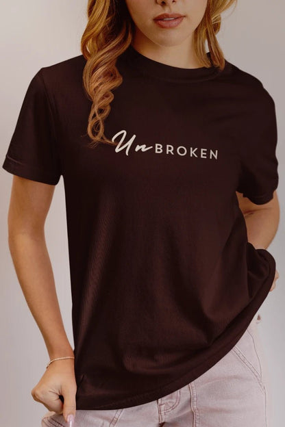 A woman wearing a coffee brown short-sleeve t-shirt with 'UnbROKEN' printed in white across the chest, paired with light pink jeans against a neutral studio background.