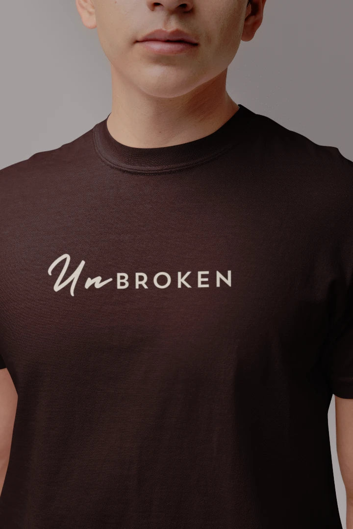 A close-up of a person wearing a coffee brown crew-neck t-shirt with the words 'Un-BROKEN' printed in white across the chest.