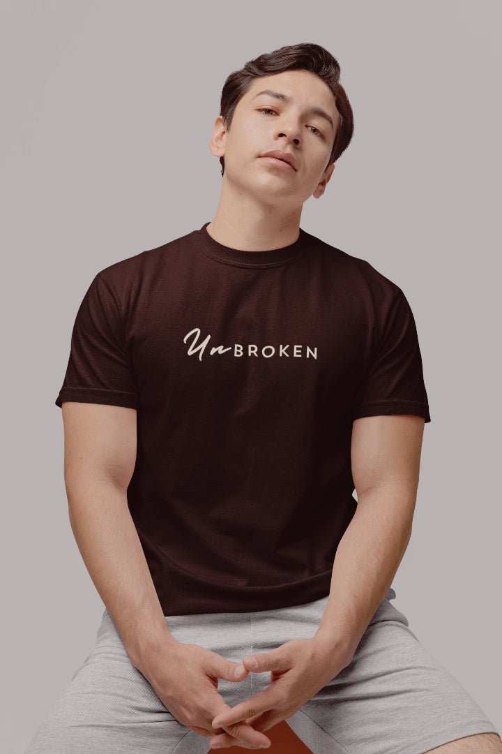 A man wearing a coffee brown t-shirt with 'Unbroken' printed in white script across the chest, paired with light gray shorts, set against a solid gray background.