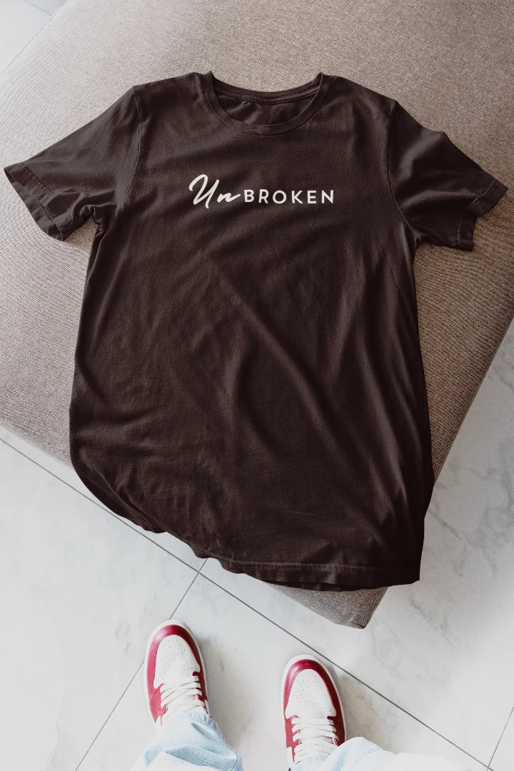 A coffee brown t-shirt with the words 'UnBROKEN' printed in white across the chest, laid flat on a gray upholstered bench with a person's feet in red and white sneakers visible at the bottom.